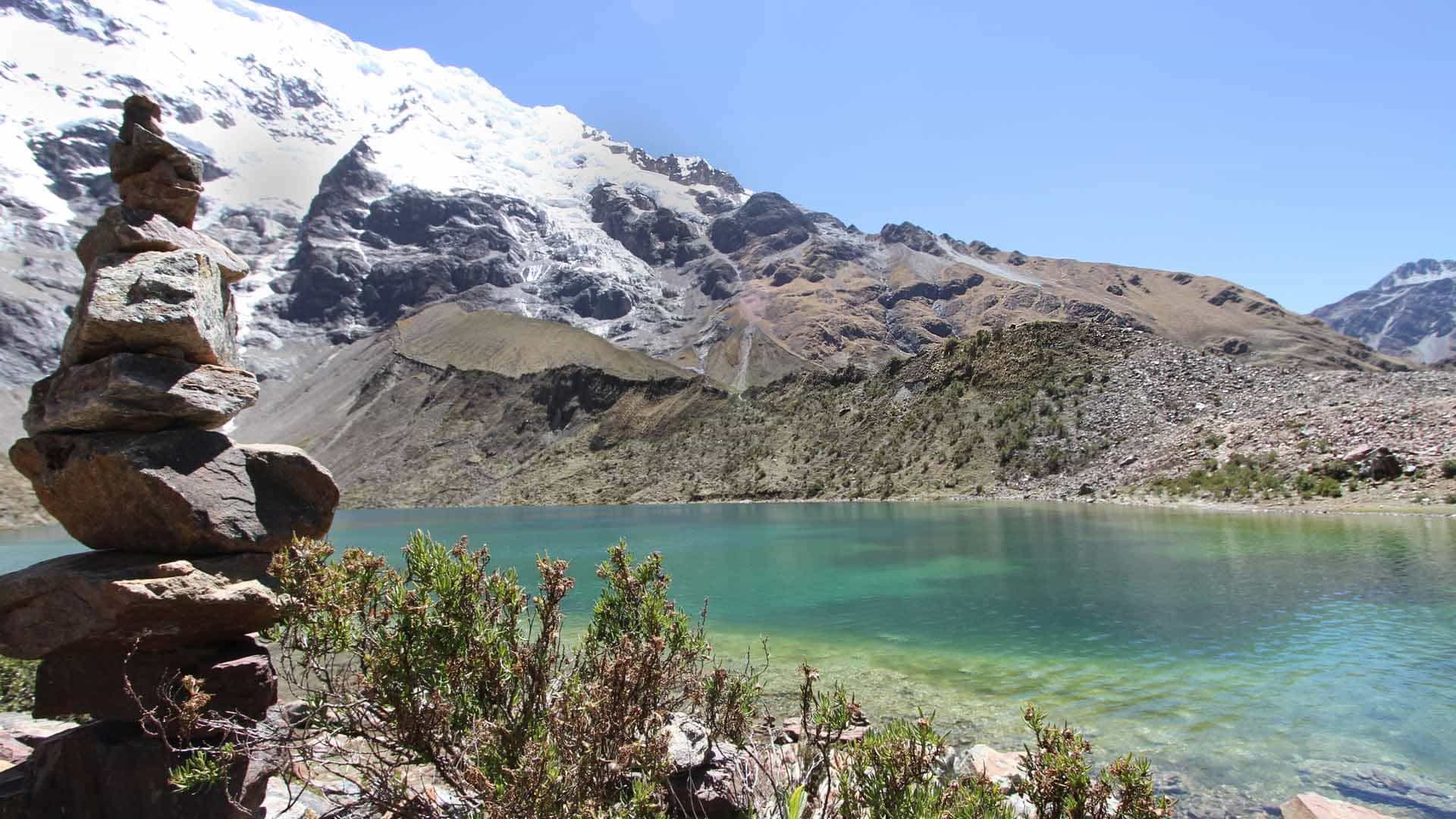 Apacheta stone cairn stacked above a turquoise glacial lake beneath the snow and ice of the Salkantay massif